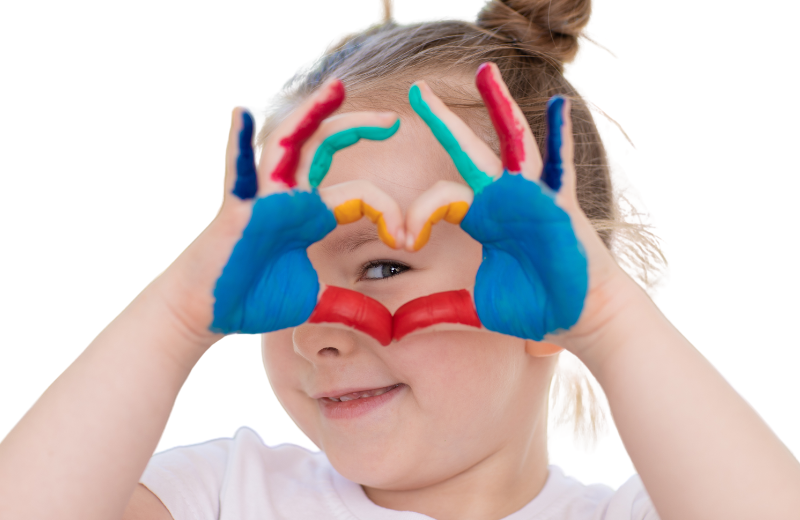 A young girl with light brown hair tied in a bun smiles playfully while making a heart shape with her painted hands. Her hands are covered in bright blue, red, green, and yellow paint, and she peeks through the heart shape she created with her fingers. The background is a clean white, making the vibrant colors of the paint stand out.